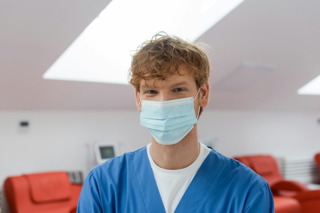 redhead and young doctor in medical mask and blue uniform looking at camera in sterile environment